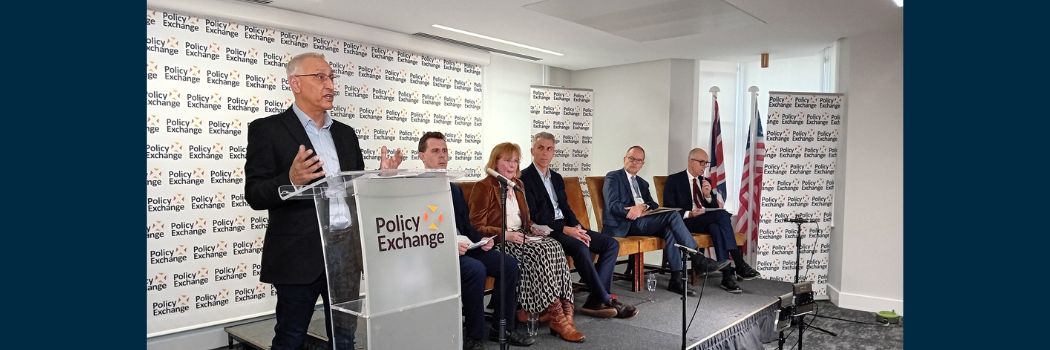 A male to the left of the picture speaks to an audience, off picture, from behind a lectern. Four others are seated in the background