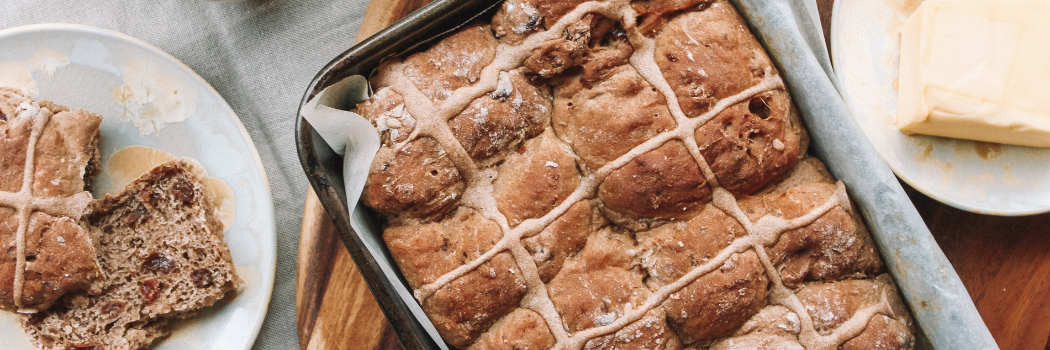 Hot cross buns on baking tray on top of table beside plate of butter