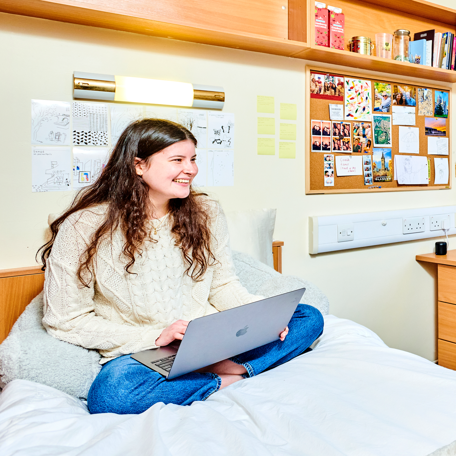 A student sits cross-legged on a bed, holding a laptop