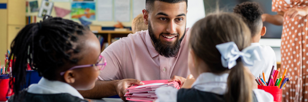 A male teacher smiling at two primary school pupils sitting at a desk in a classroom