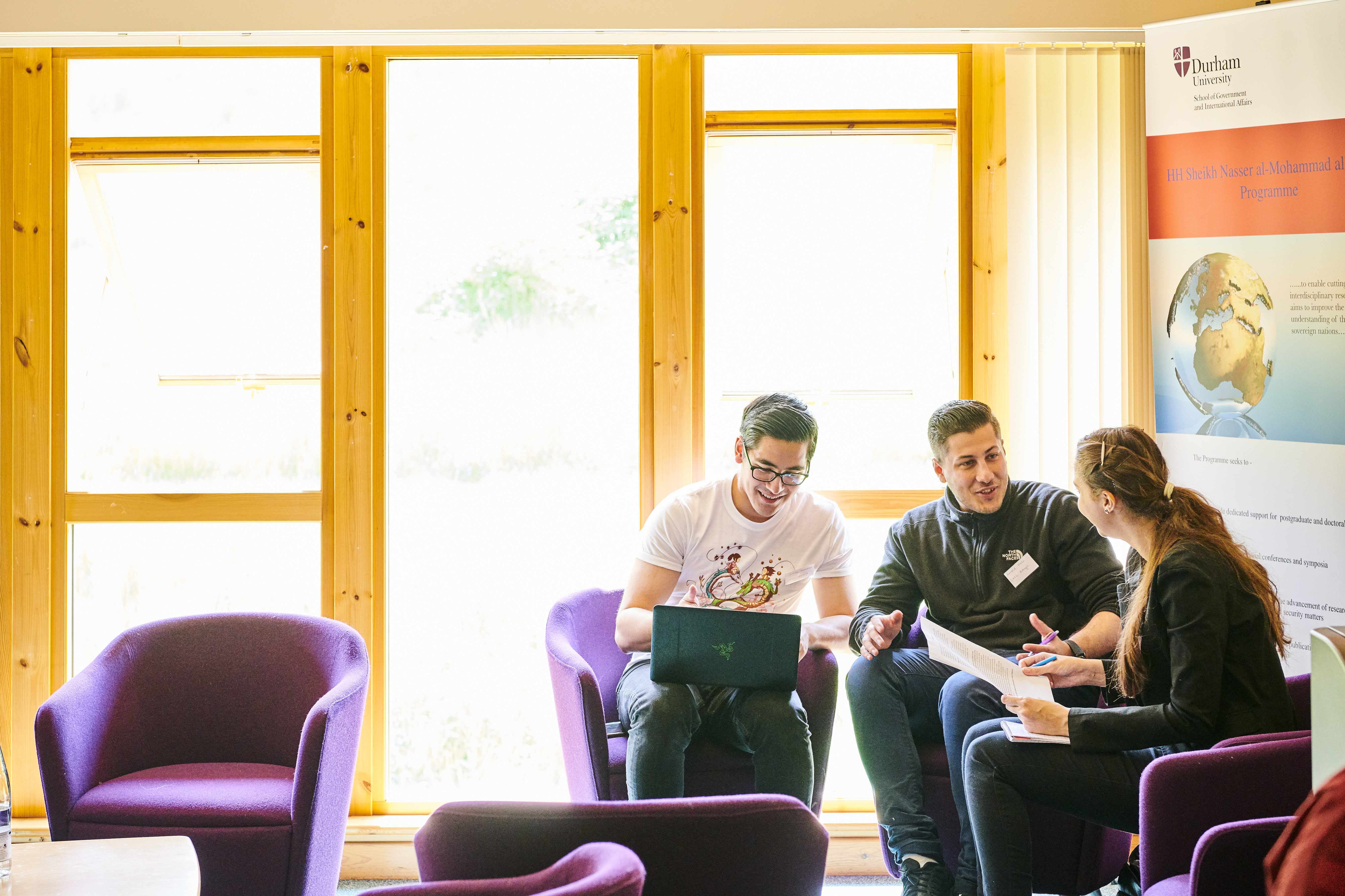 TThree students sitting in a sunlit common room having a discussion