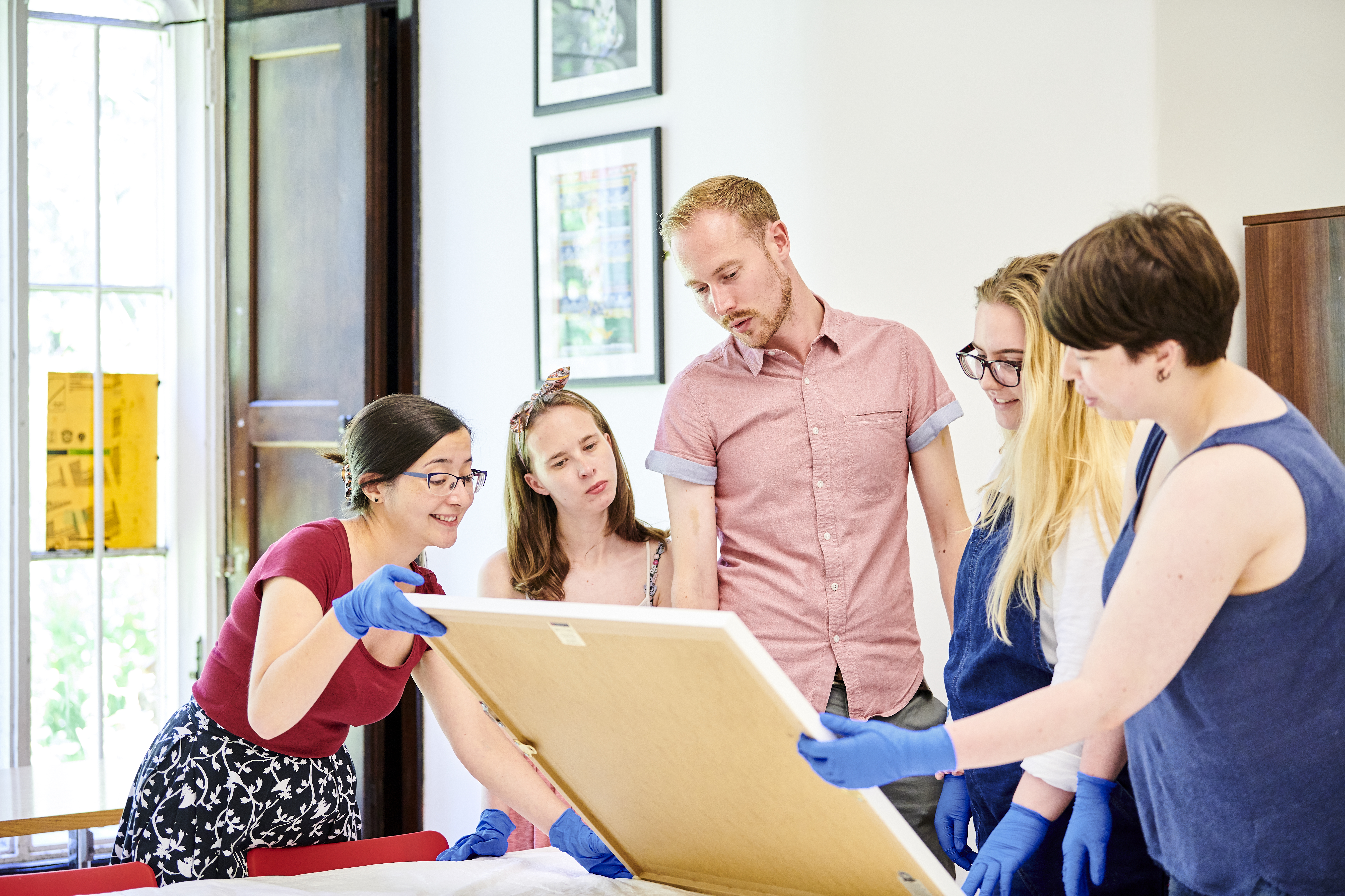 A group of people wearing gloves stand around an artwork propped up on a table having a discussion