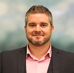 A smiling man with a close-cut beard wearing a light pink shirt and jacket