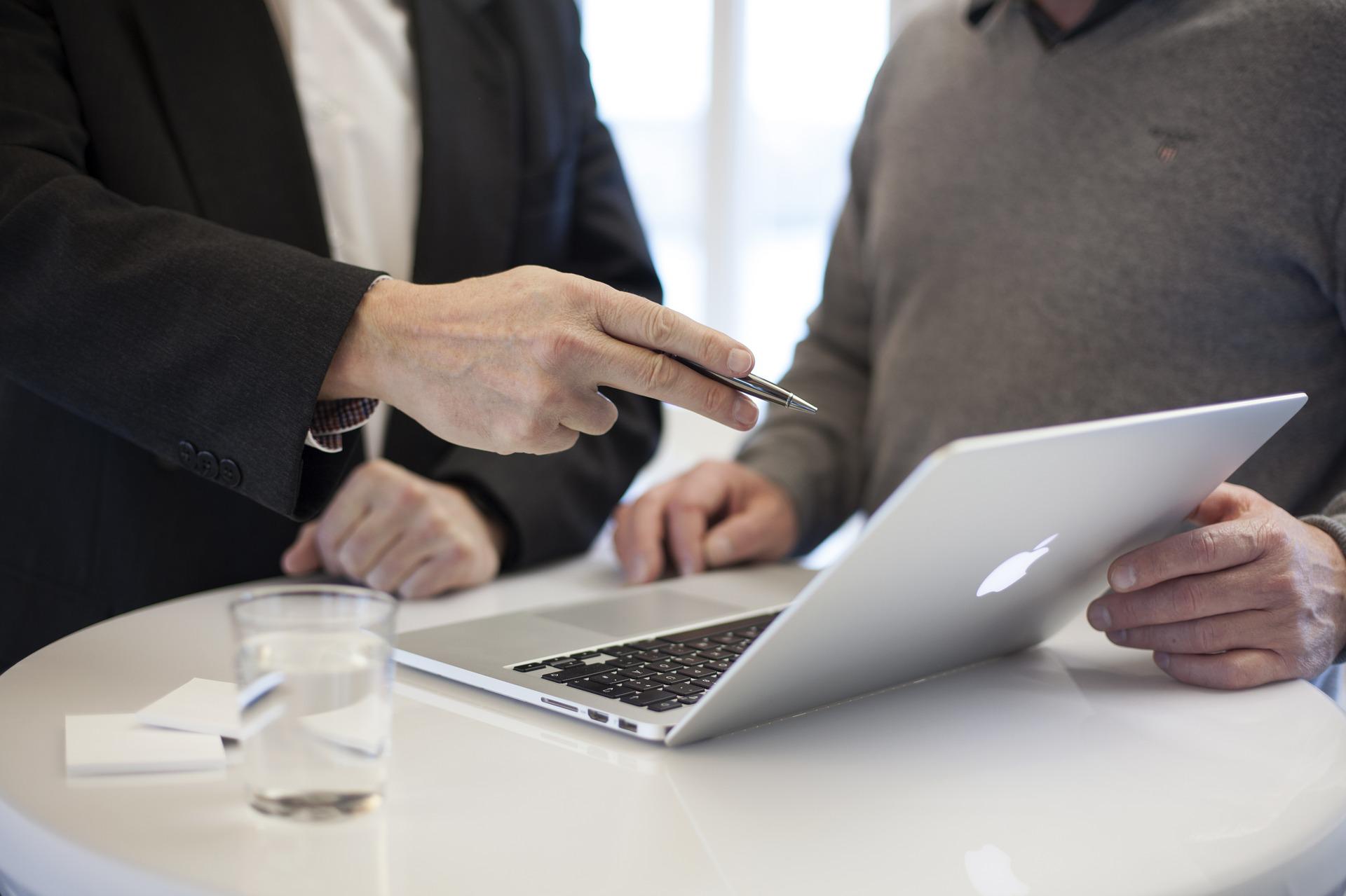 two people sitting down with a laptop between them and a glass of water on the table. One of them is pointing toward the screen with a pen