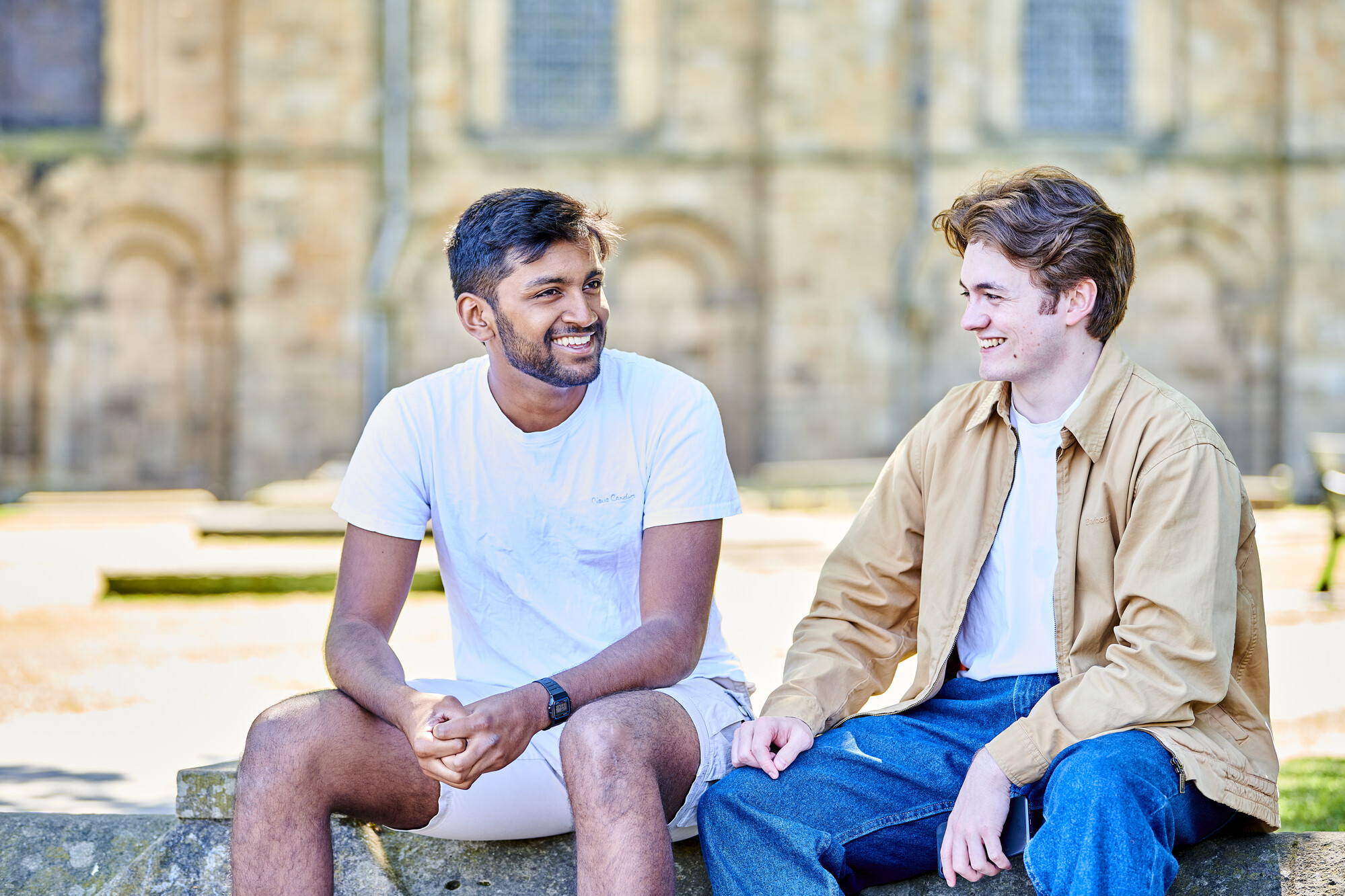 Students dressed in casual clothes sit / stand in front of the Cathedral which is visible in the background - it is a sunny day and they all look happy